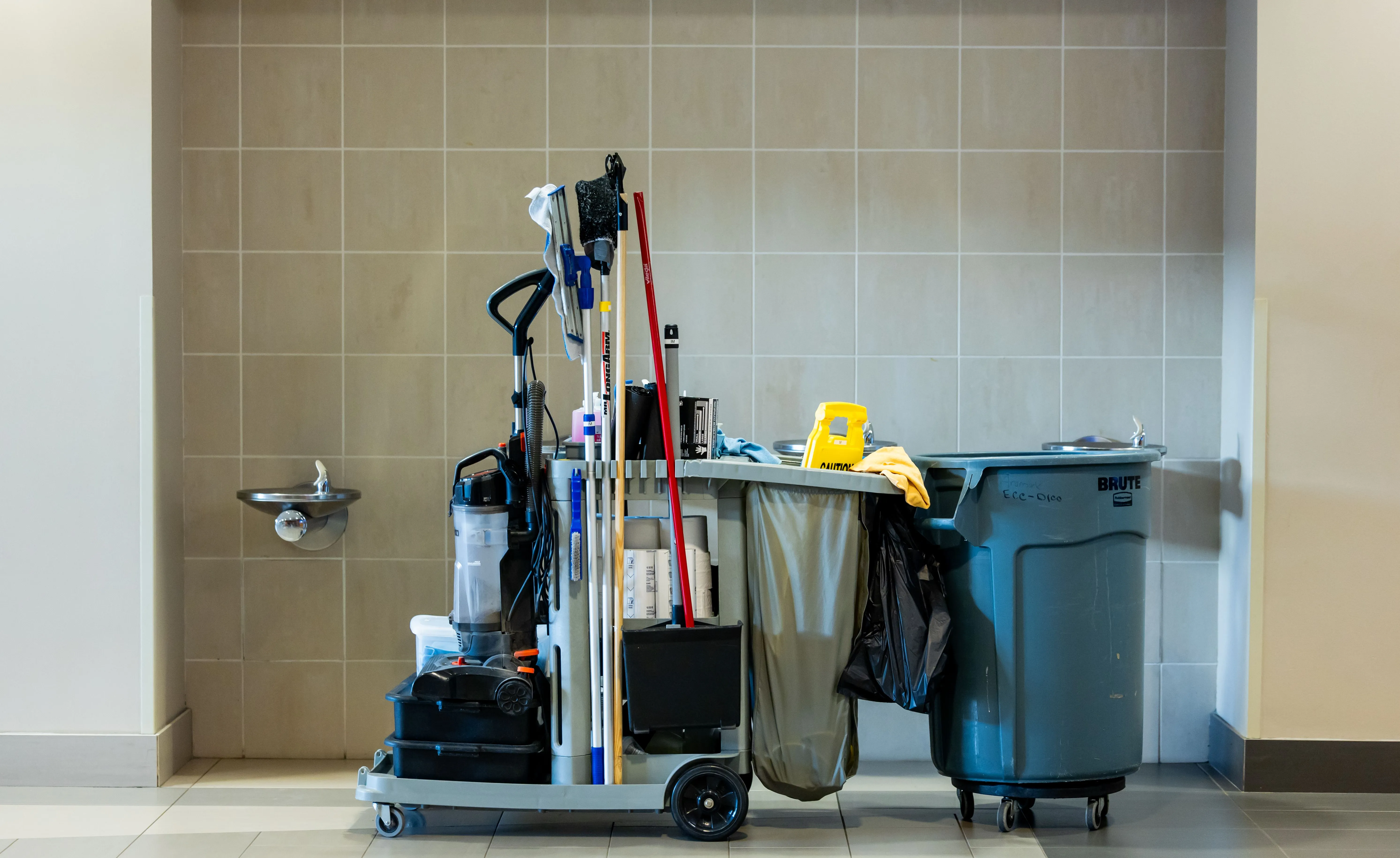 Keep clean - Reinigungs - Ein voll beladener Reinigungswagen steht vor einer gefliesten Wand mit einem Trinkbrunnen.