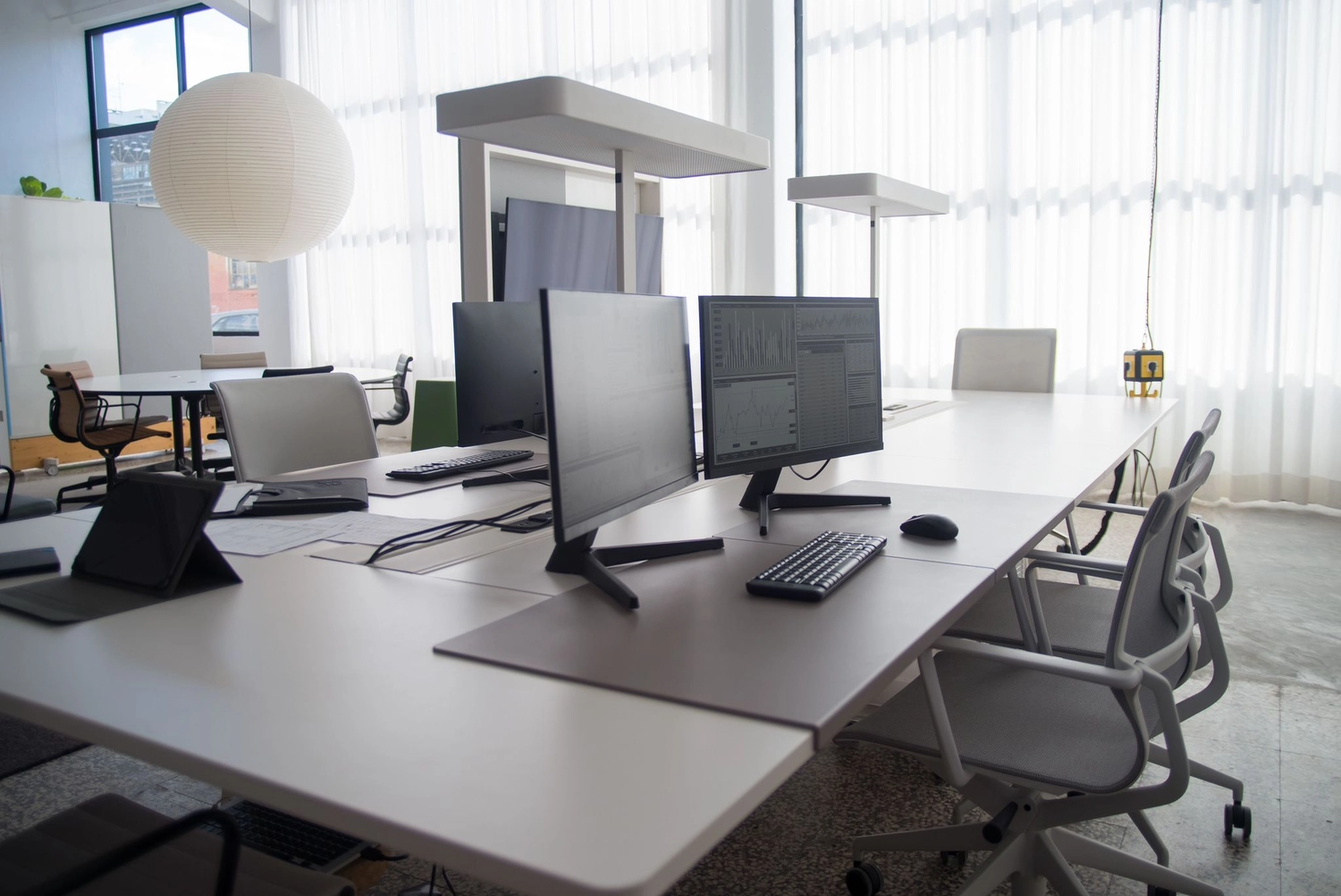 Keep clean - Reinigungs - Moderner Büroarbeitsplatz mit Reihen langer weißer Schreibtische, mehreren Computermonitoren, Tastaturen und ergonomischen Stühlen vor großen, mit Vorhängen versehenen Fenstern.
